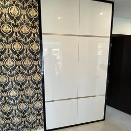 STOKŁOSA - Meble na wymiar RAFAŁ STOKŁOSA - Biała szafa przesuwna z czarną ramą, podzielona poziomymi paskami, na tle ściany w ciemny wzorzysty deseń. Widoczny fragment podłogi z jasnych płytek.