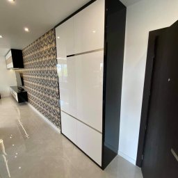 STOKŁOSA - Meble na wymiar RAFAŁ STOKŁOSA - Nowoczesna szafa wnękowa z białymi frontami i czarnymi bokami, dopasowana do wzorzystej tapety w salonie. Błyszcząca podłoga odbija światło, podkreślając elegancję mebla.