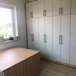 STOKŁOSA - Meble na wymiar RAFAŁ STOKŁOSA - Jasna szafa wnękowa z czarnymi uchwytami i biurko z drewnianym blatem w pokoju z oknem. Widoczna podłoga imitująca drewno i logo firmy na ścianie.