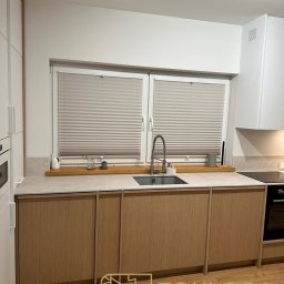 STOKŁOSA - Meble na wymiar RAFAŁ STOKŁOSA - Jasna kuchnia z drewnianymi frontami szafek, szarym blatem i oknem z plisowanymi roletami. Minimalistyczny styl, nowoczesne wykończenie, widoczny zlew i bateria.