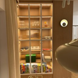 STOKŁOSA - Meble na wymiar RAFAŁ STOKŁOSA - Biała, podświetlana szafa z ryflowanymi szklanymi drzwiami, wypełniona przedmiotami. Obok lampa ścienna i zasłona. Ujęcie z bliska, ciepłe światło.