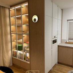 STOKŁOSA - Meble na wymiar RAFAŁ STOKŁOSA - Nowoczesna kuchnia na wymiar z przeszkloną witryną, oświetleniem punktowym i białymi frontami. Widoczny fragment jadalni i logo firmy Tokłosa.