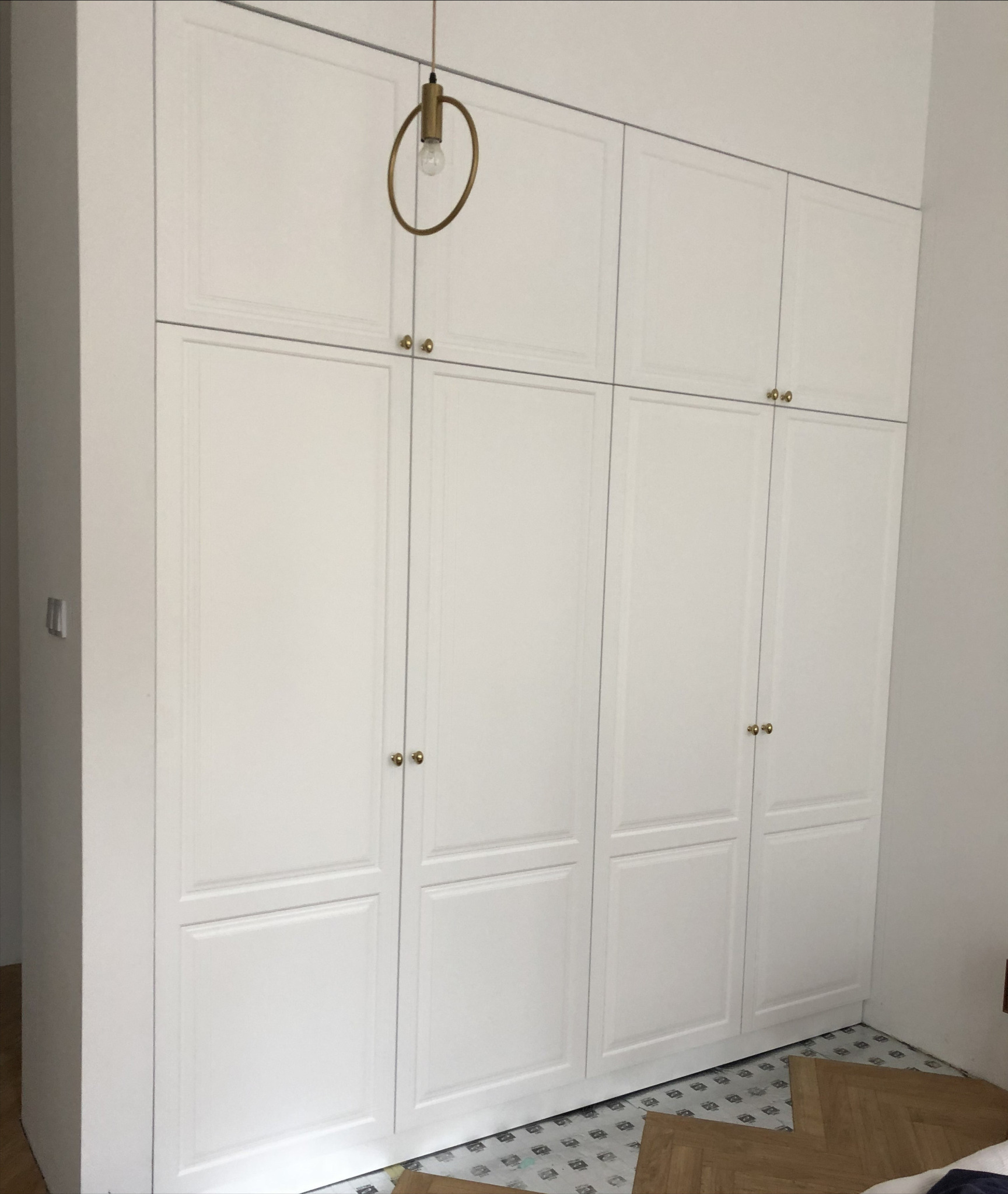 Biała, zabudowana szafa z górnymi szafkami, złotymi uchwytami i okrągłą lampą wiszącą, widoczna na tle fragmentu podłogi z wzorem geometrycznym.