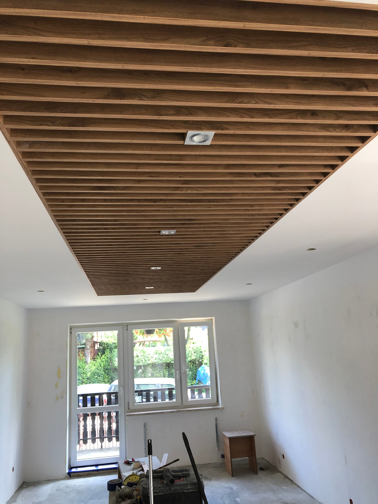 Drewniany sufit lamelowy z wbudowanym oświetleniem punktowym w trakcie montażu, widoczne narzędzia i niedokończone ściany w pomieszczeniu.