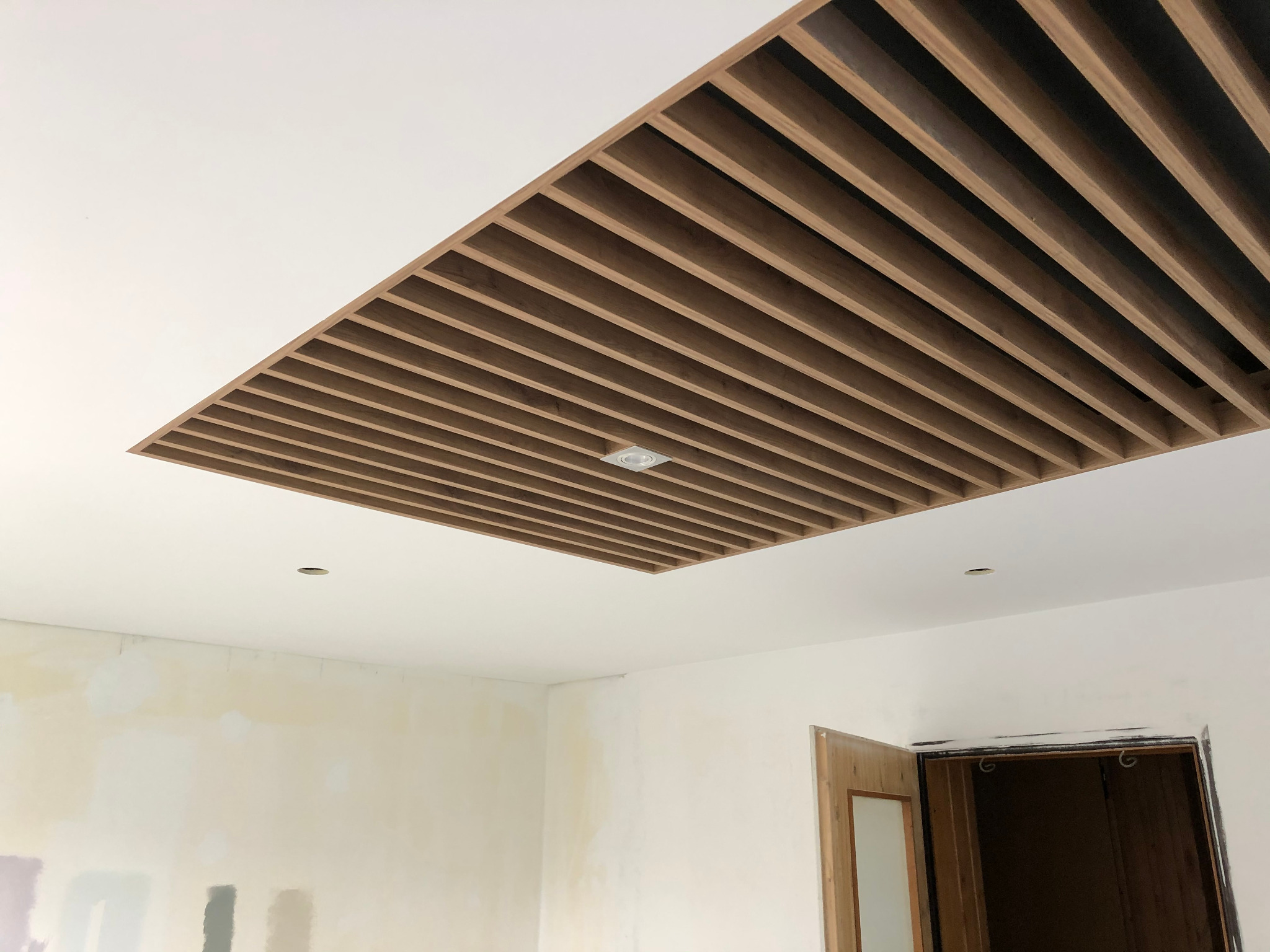 Drewniana konstrukcja sufitowa z listew w prostokątnej wnęce, widoczna instalacja oświetleniowa, surowe ściany w trakcie wykończenia, otwarte drzwi do innego pomieszczenia.