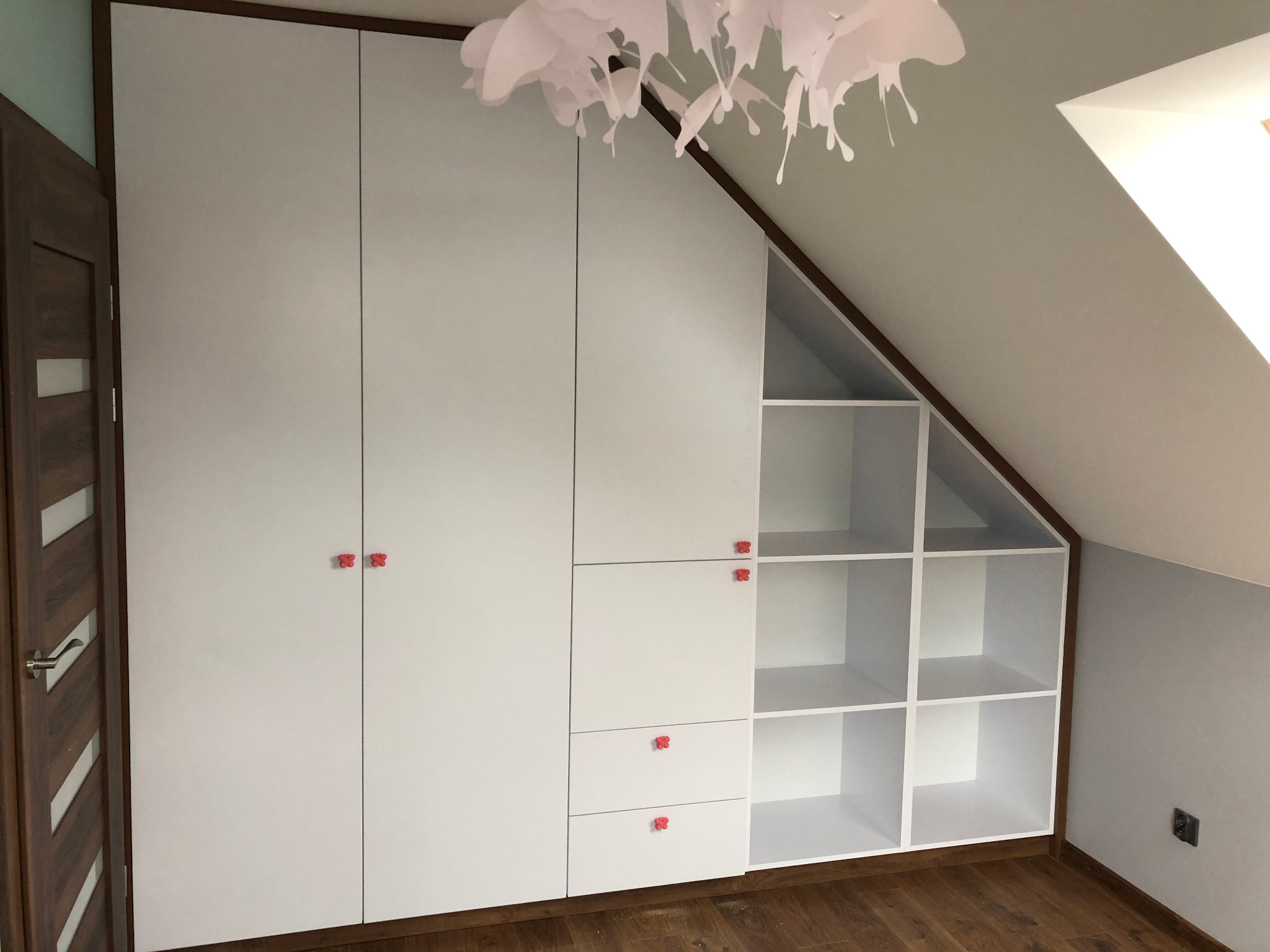 Biała szafa wnękowa z czerwonymi uchwytami w kształcie kwiatków, dopasowana do skosu poddasza, z otwartymi półkami i szufladami, oświetlona dekoracyjną lampą.