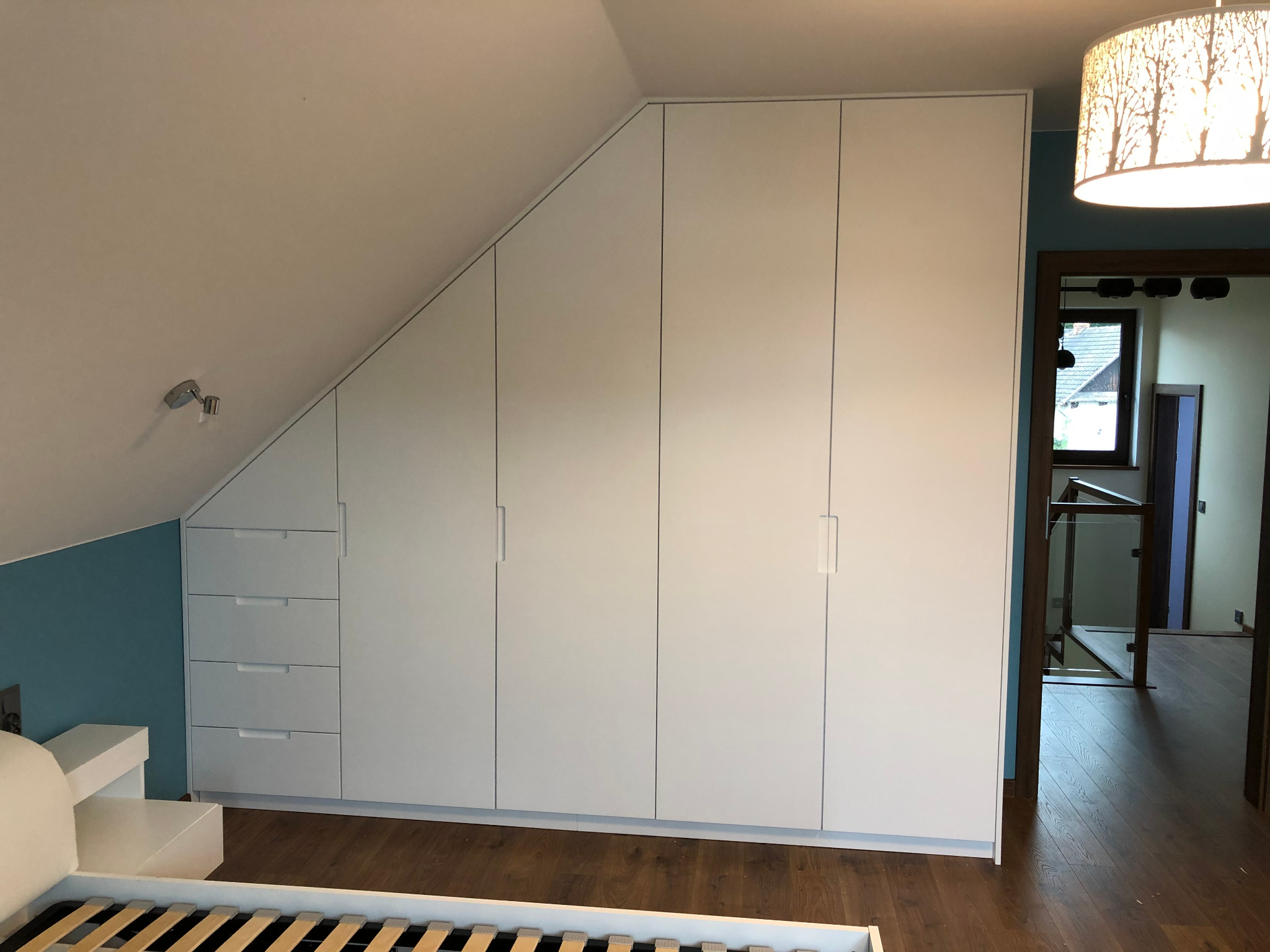 Biała szafa wnękowa z szufladami dopasowana do skosu dachu w sypialni z turkusową ścianą akcentującą.