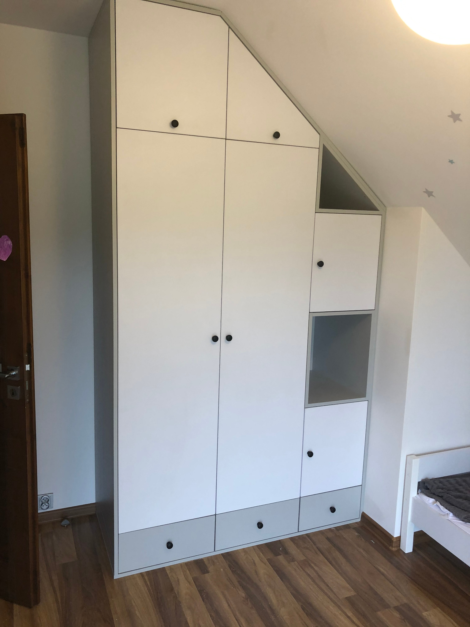 Biała szafa wnękowa na wymiar z szarymi akcentami, dopasowana do skosu dachu w pokoju dziecięcym, z widocznym łóżkiem i naklejkami gwiazd na suficie.