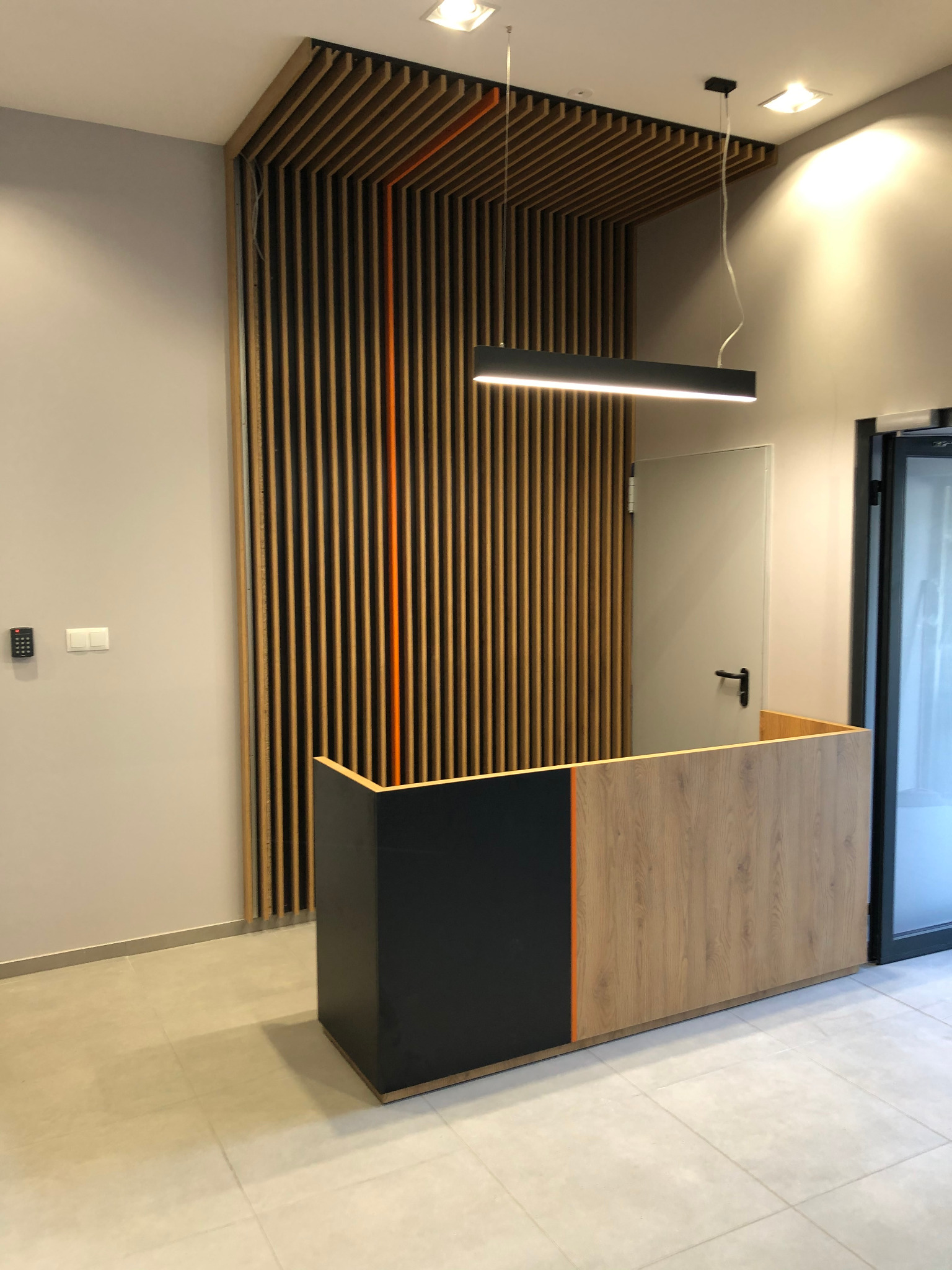 Recepcja z drewnianymi lamelami na ścianie, oświetlona podłużną lampą, z elementami w kolorze czarnym i drewnopodobnym.