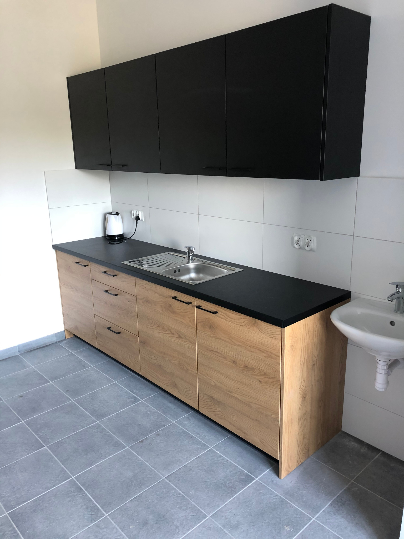Nowoczesna kuchnia: dolne szafki z drewnopodobnym frontem i czarnym blatem, górne szafki czarne, bateria kuchenna, czajnik elektryczny na blacie, płytki ścienne i podłogowe w odcieniach szarości.