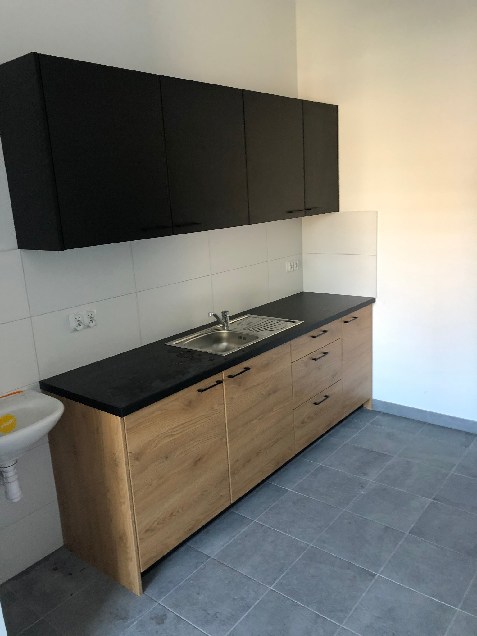 Nowoczesna kuchnia: górne szafki czarne, dolne z drewnianym fornirem, czarny blat i stalowy zlew, białe płytki na ścianie.