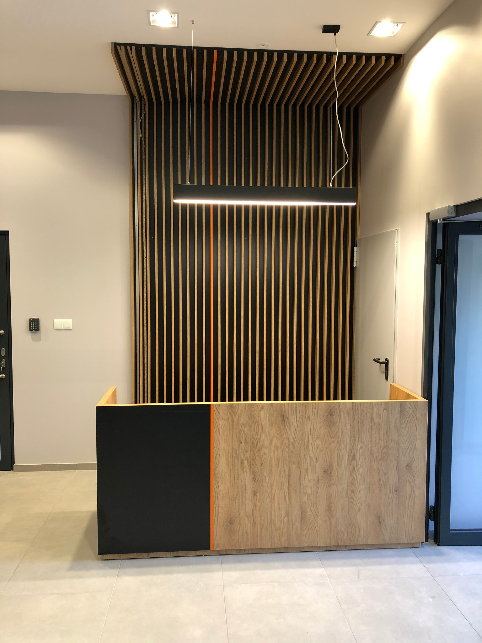 Recepcja z drewnianym blatem i czarnym panelem, oświetlona wiszącą lampą, z dekoracyjną ścianą z pionowych listew w tle.