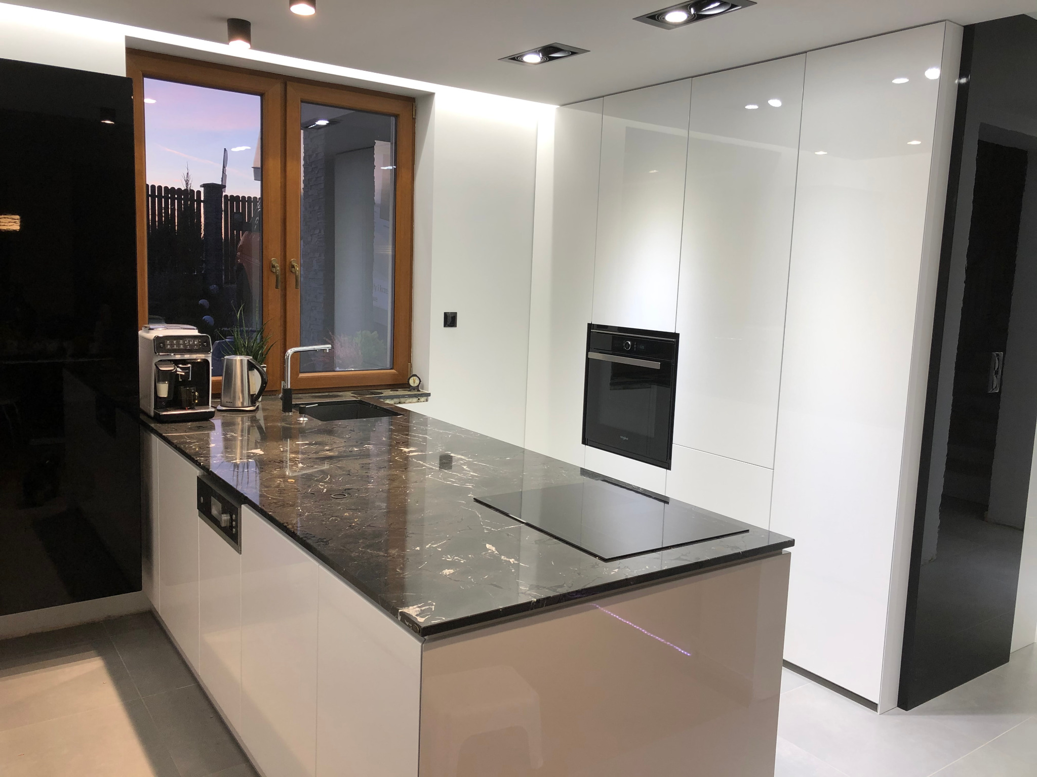 Nowoczesna kuchnia: białe, lakierowane fronty szafek, ciemny blat z marmurkowym wzorem, okno z widokiem na ogród i zachodzące słońce.