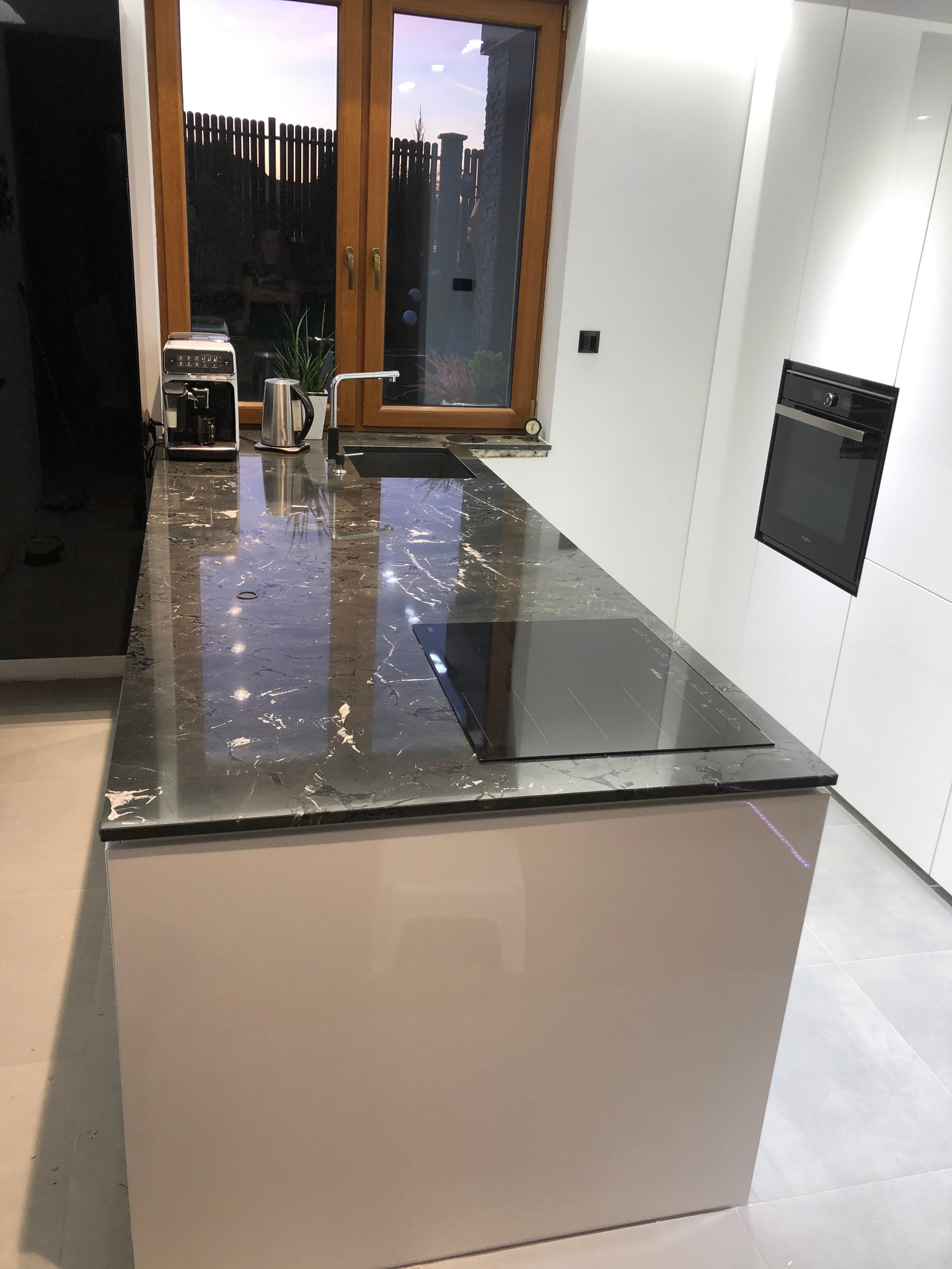 Nowoczesna kuchnia z wyspą, czarny blat z marmurowym wzorem, białe szafki i zabudowany piekarnik, ekspres do kawy na blacie przy oknie.