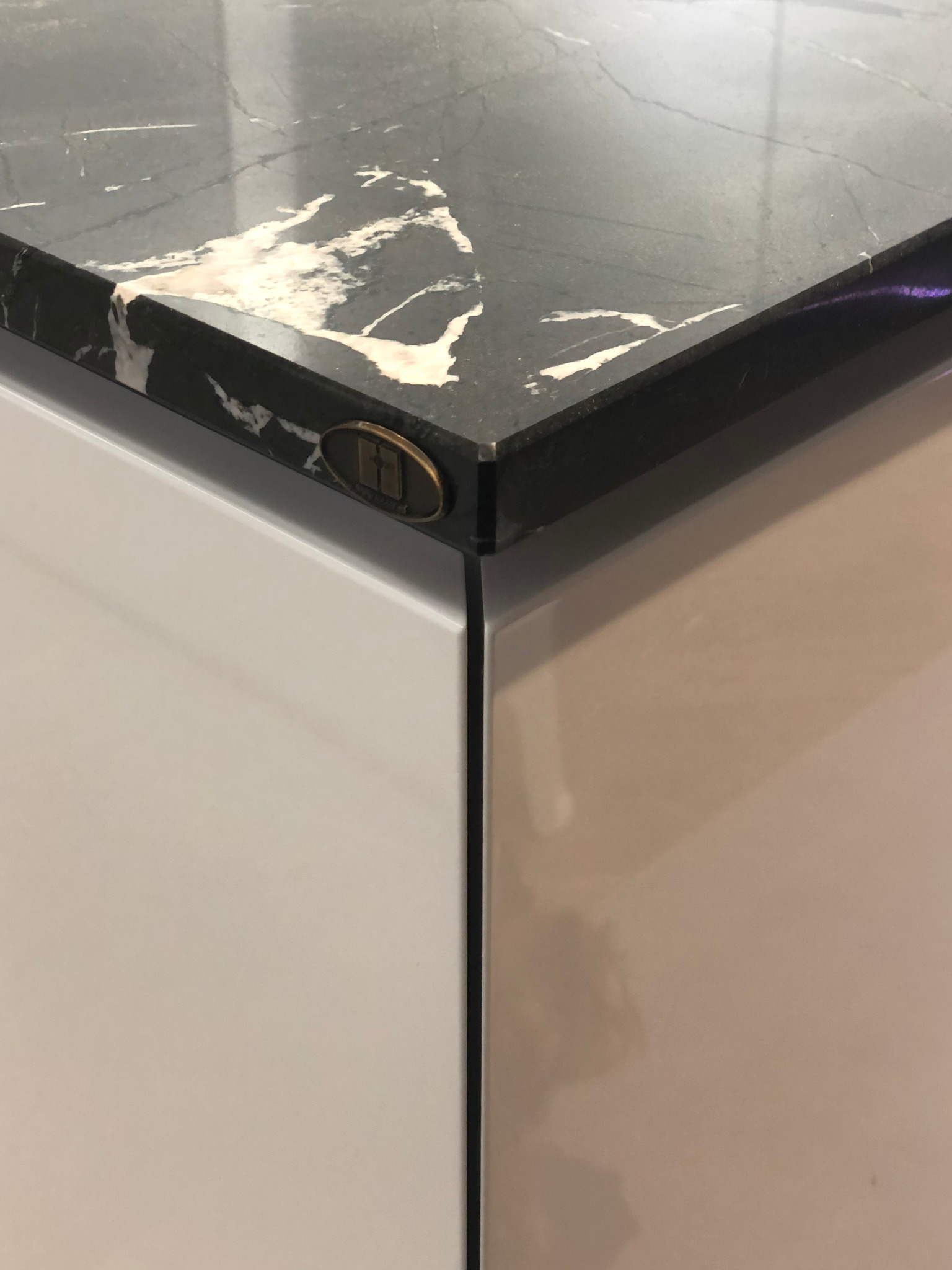 Fragment czarnego blatu z marmurowym wzorem, połączonego z białym frontem meblowym. Na krawędzi blatu widoczny okrągły, metalowy emblemat.