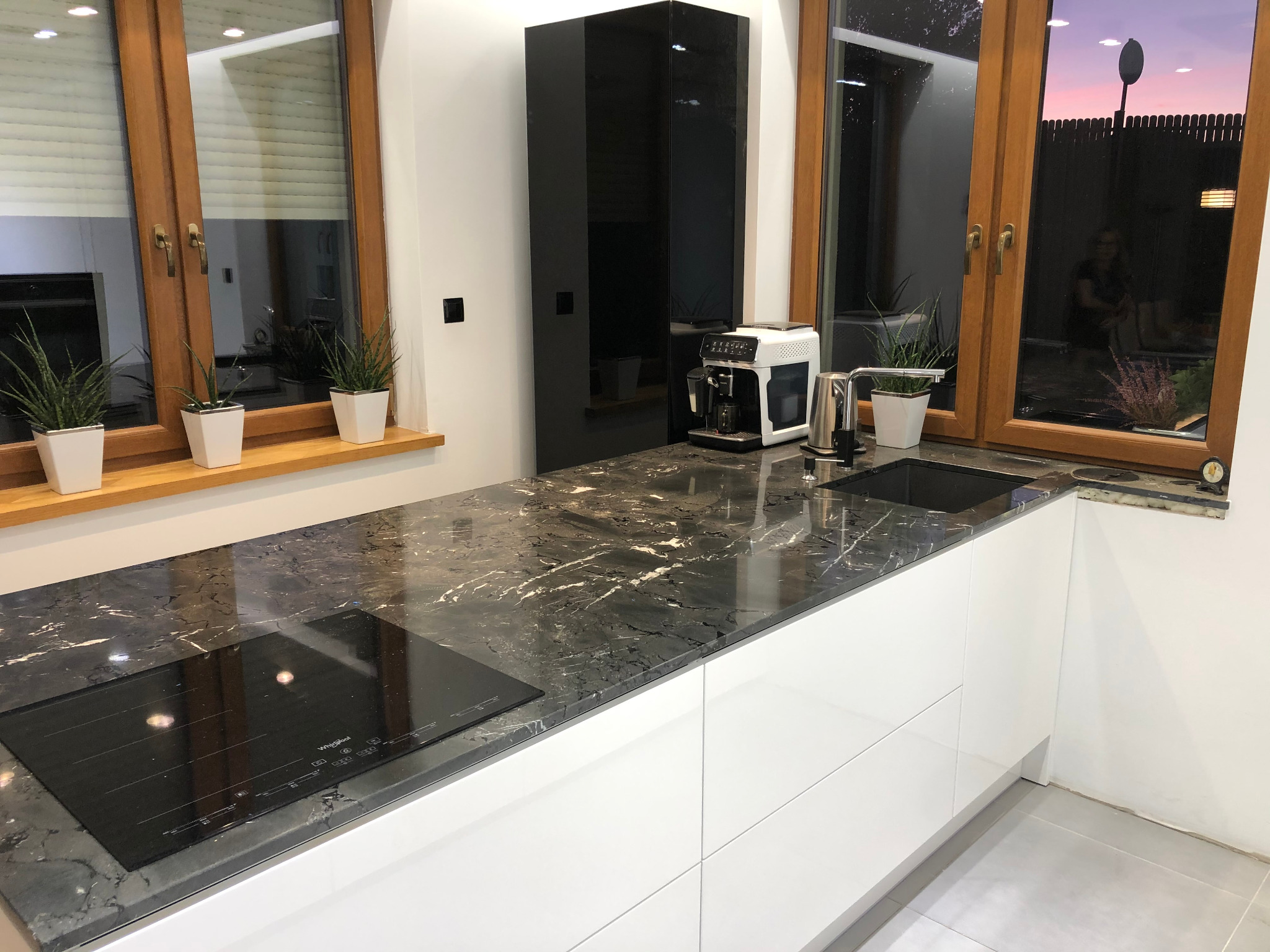 Nowoczesna kuchnia z białymi szafkami i czarnym blatem o marmurkowym wzorze, z płytą indukcyjną i ekspresem do kawy, widok na okno z roślinami doniczkowymi i zachodzącym słońcem.