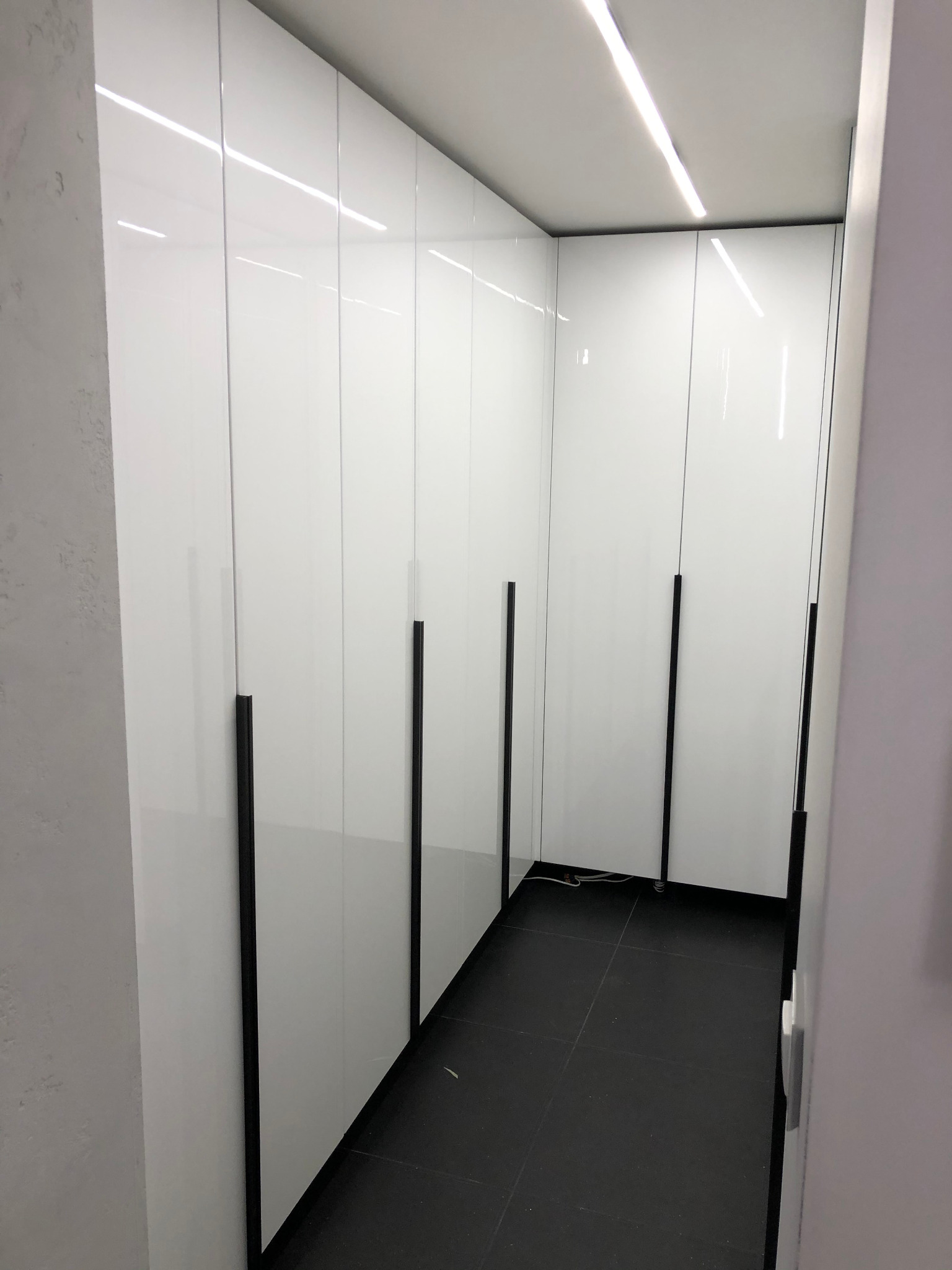 Wnętrze garderoby z białymi, lakierowanymi szafami wnękowymi z długimi, czarnymi uchwytami i czarną podłogą z płytek, oświetlone listwą LED w suficie.