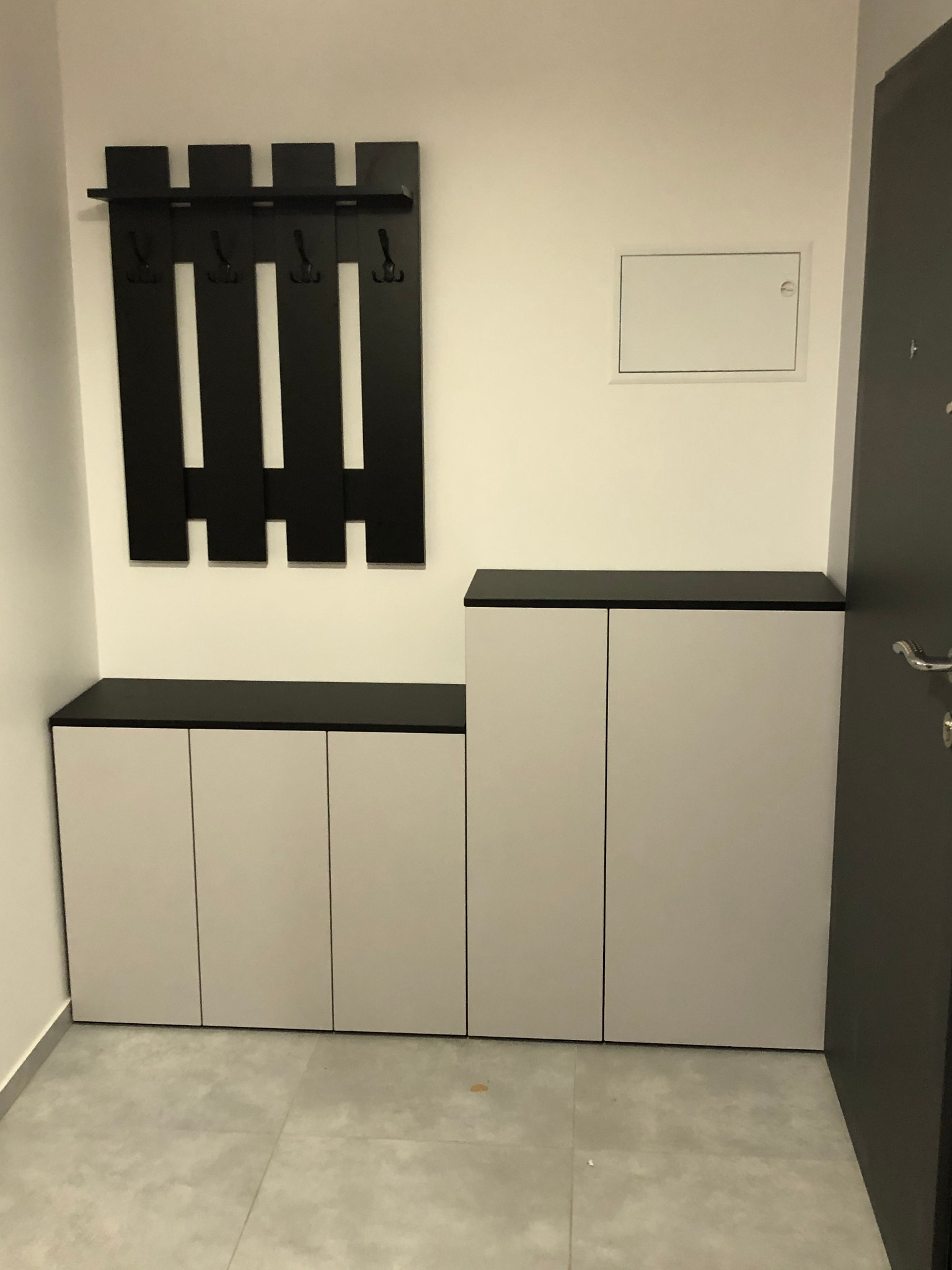 Przedpokój z zabudową: szafki z białymi frontami i czarnym blatem, czarny wieszak na ścianie z półką i haczykami, szara podłoga z płytek.