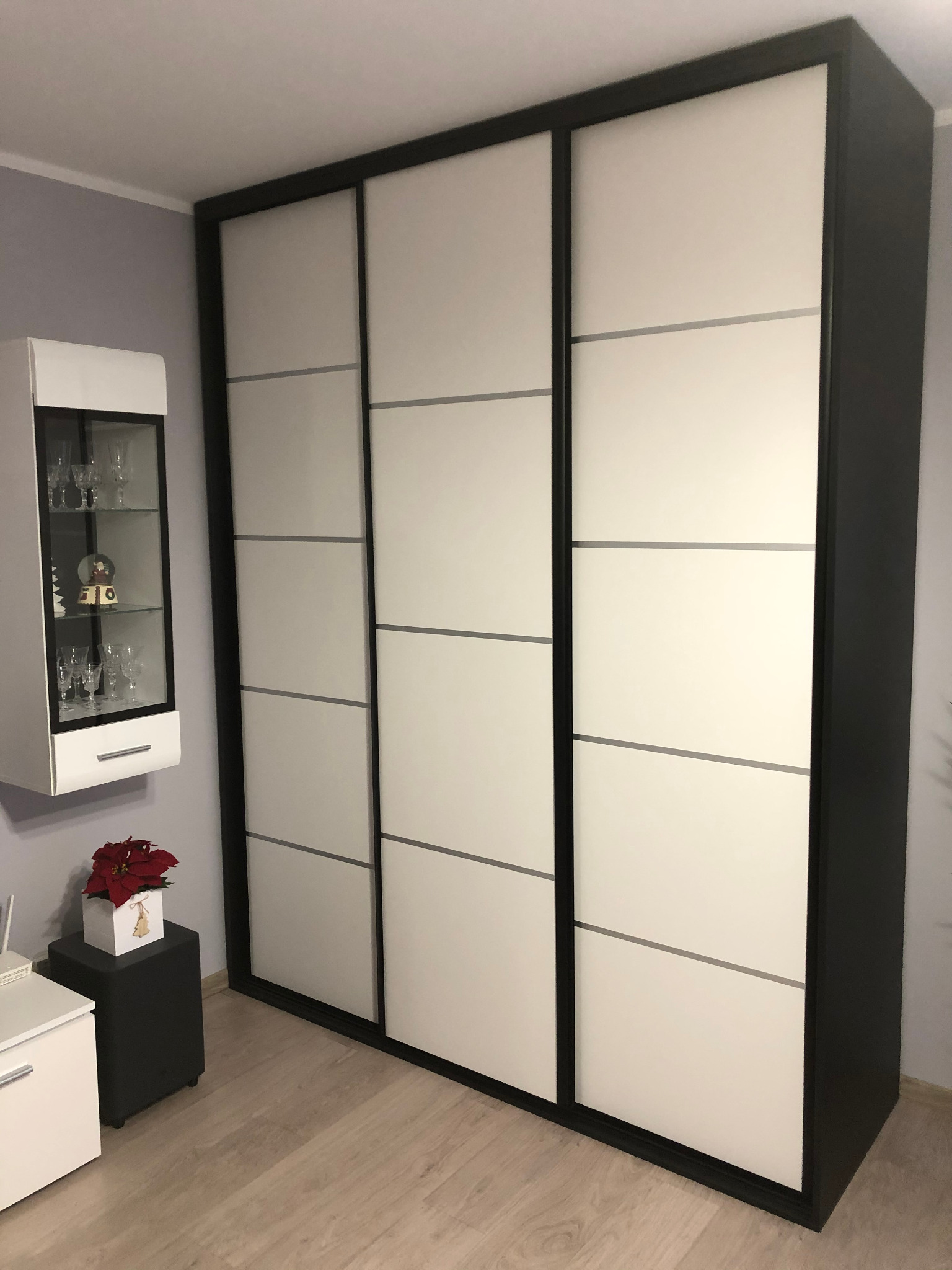 Duża szafa przesuwna z trzema białymi panelami i czarną ramą, widoczna w rogu pokoju z szarą ścianą i drewnianą podłogą. Obok szafy stoi witryna z szklanymi drzwiami i dekoracjami świątecznymi.