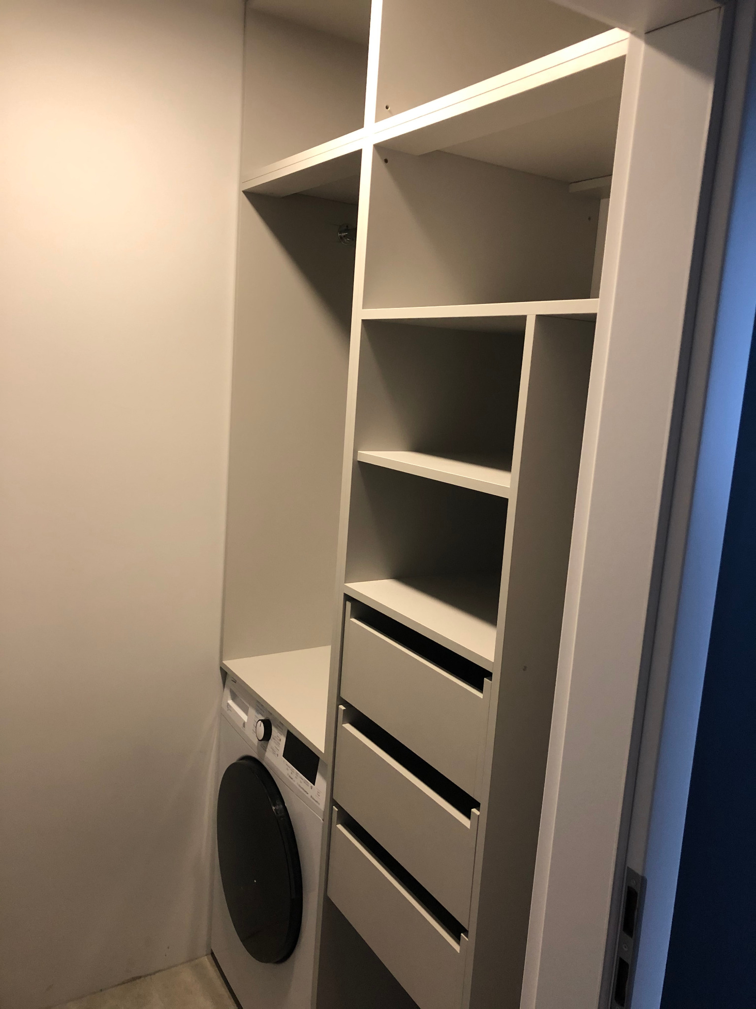 Biała szafa wnękowa z szufladami i półkami, obok pralka w łazience.
