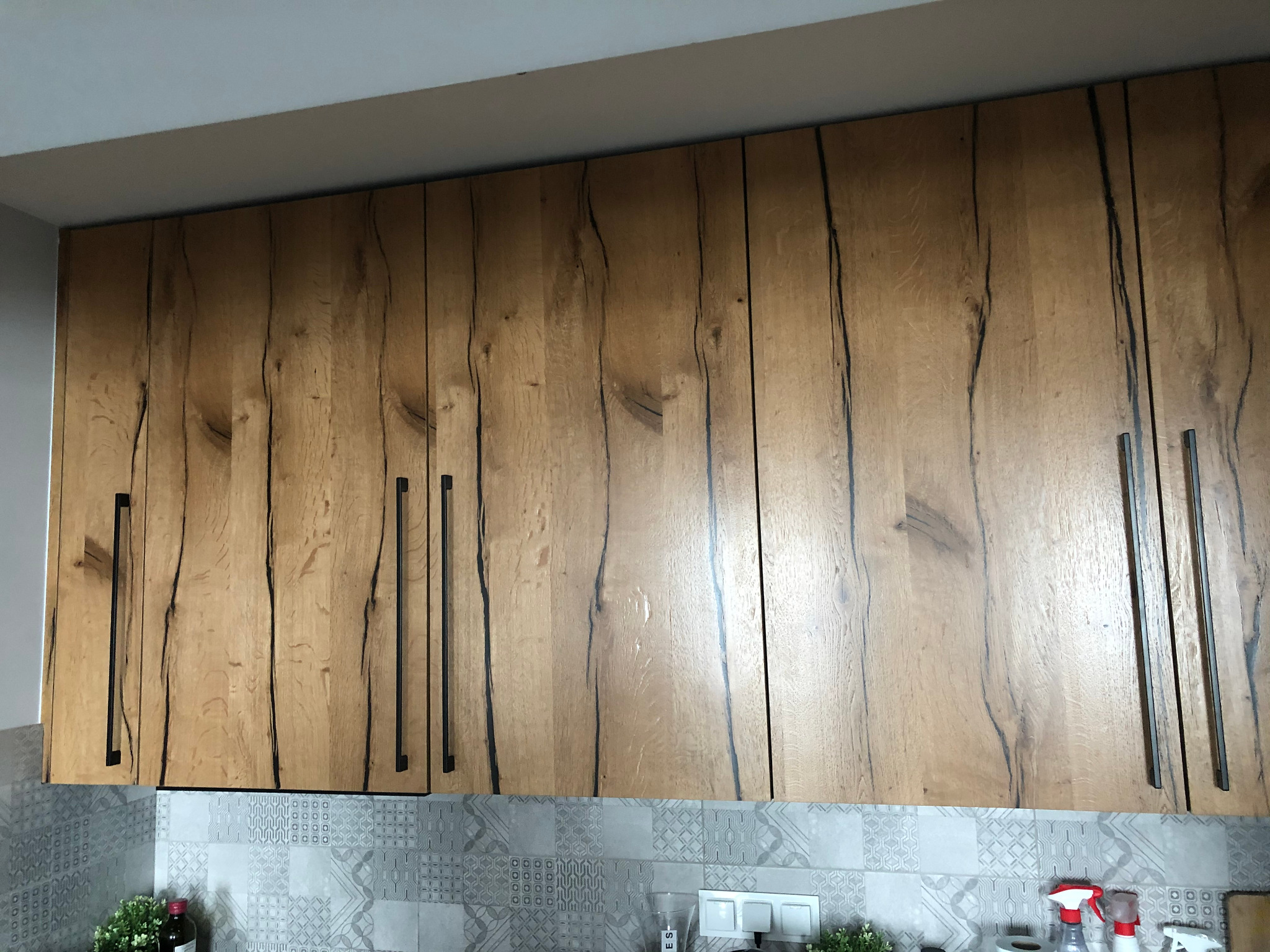 Górny rząd szafek kuchennych z drewnianymi frontami o wyraźnym usłojeniu i długich, czarnych uchwytach, na tle mozaiki ściennej.