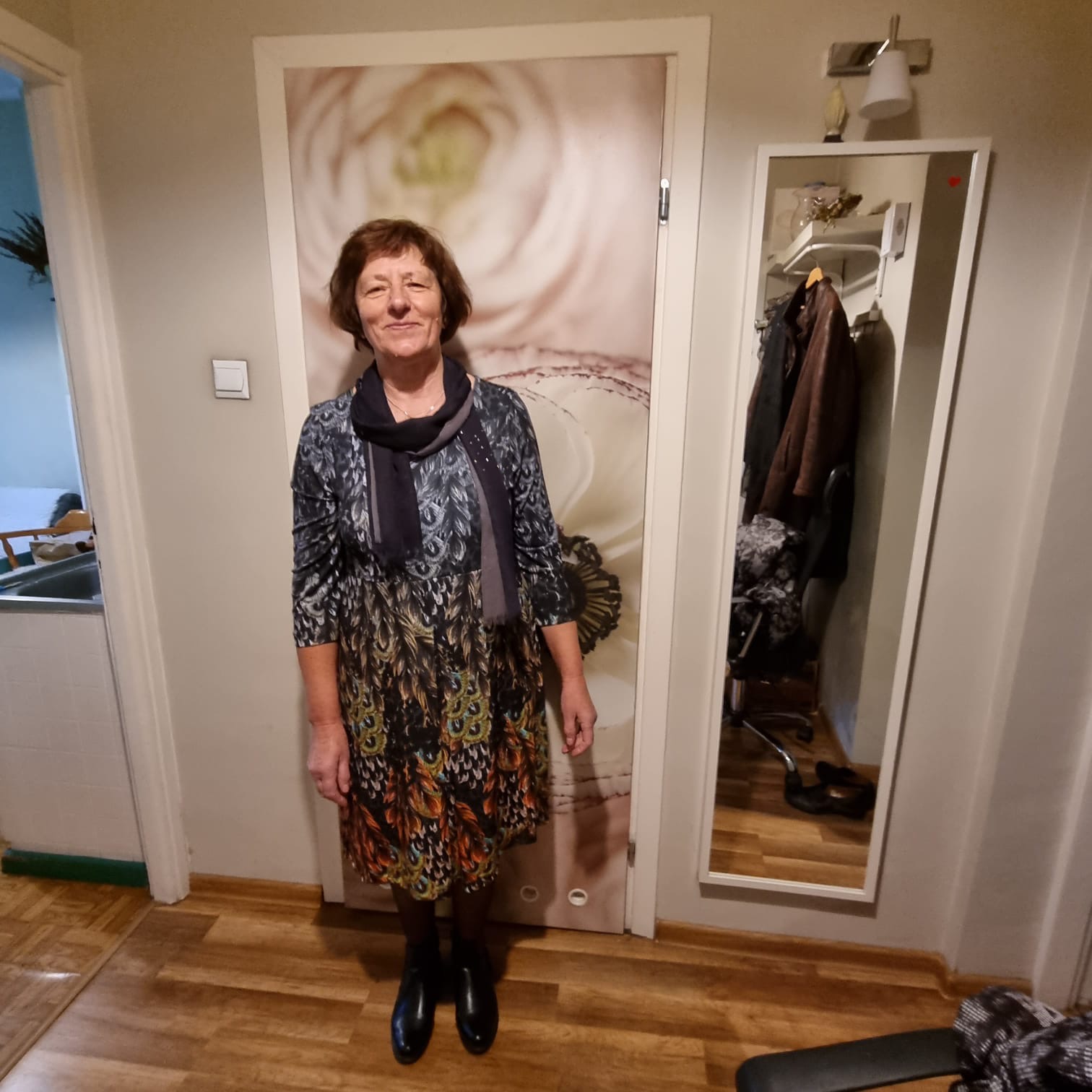 Kobieta w kwiecistej sukience i czarnych butach stoi w korytarzu przed drzwiami z fototapetą kwiatu, obok lustra z widoczną zawartością szafy.