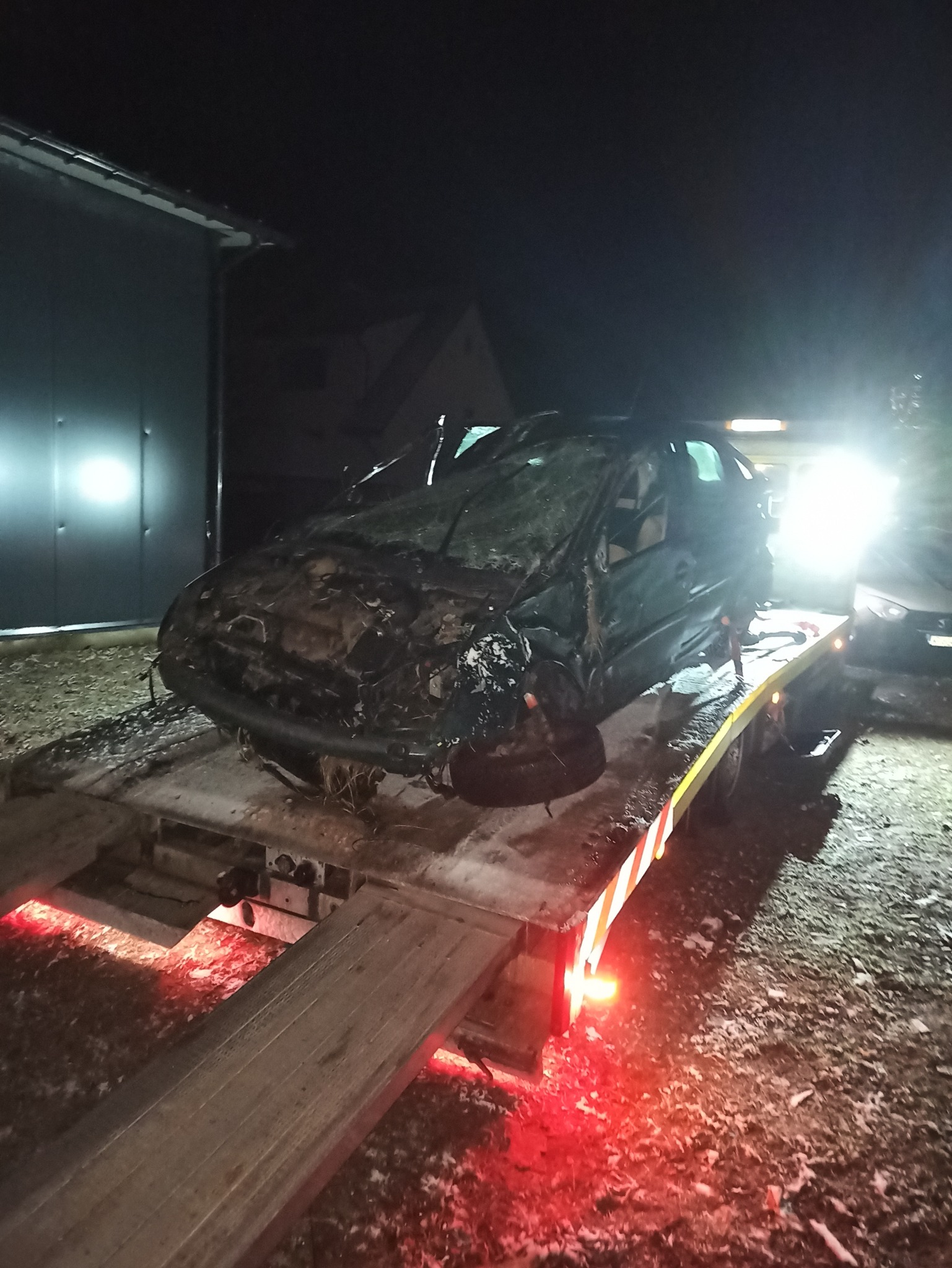 Uszkodzony samochód osobowy, z widocznymi zniszczeniami karoserii i wybitymi szybami, transportowany na platformie lawety w warunkach nocnych, oświetlony przez reflektory i czerwone światła...