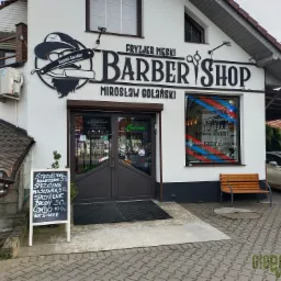 Fasada budynku z szyldem 'Fryzjer Męski Barber Shop Mirosław Golański', logo z brzytwą i kapeluszem, okno z naklejkami reklamowymi oraz tablica z cennikiem usług fryzjerskich.