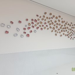 Greenwood Grzegorz Dziergas - Dekoracyjna instalacja ścienna z trójwymiarowych elementów w kształcie płatków, stopniowo zmieniających kolor od srebrnego i różowego do brązowego, na białej ścianie z dolnym pasem mozaiki.