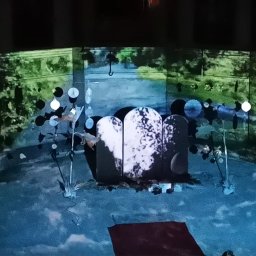 Greenwood Grzegorz Dziergas - Scenografia teatralna z projekcjami: stylizowany ogród z nagrobkami w tle, oświetlony dekoracyjnymi lampkami i z elementami roślinnymi.