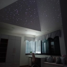 Greenwood Grzegorz Dziergas - Pokój dziecięcy z oświetleniem sufitowym imitującym rozgwieżdżone niebo, widoczny biały regał, biurko z krzesłem i okno z zasłonami w kropki.