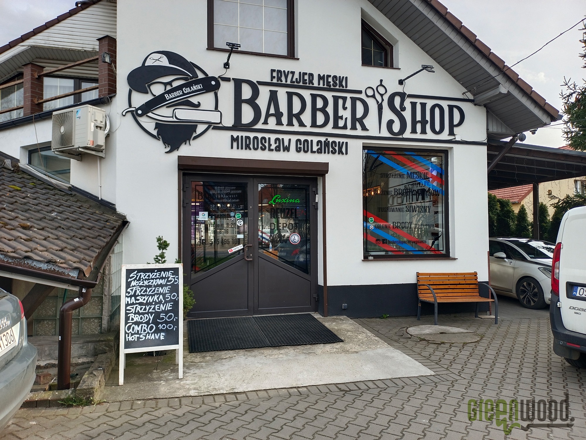 Fasada budynku z szyldem 'Fryzjer Męski Barber Shop Mirosław Golański', logo z brzytwą i kapeluszem, okno z naklejkami reklamowymi oraz tablica z cennikiem usług fryzjerskich.