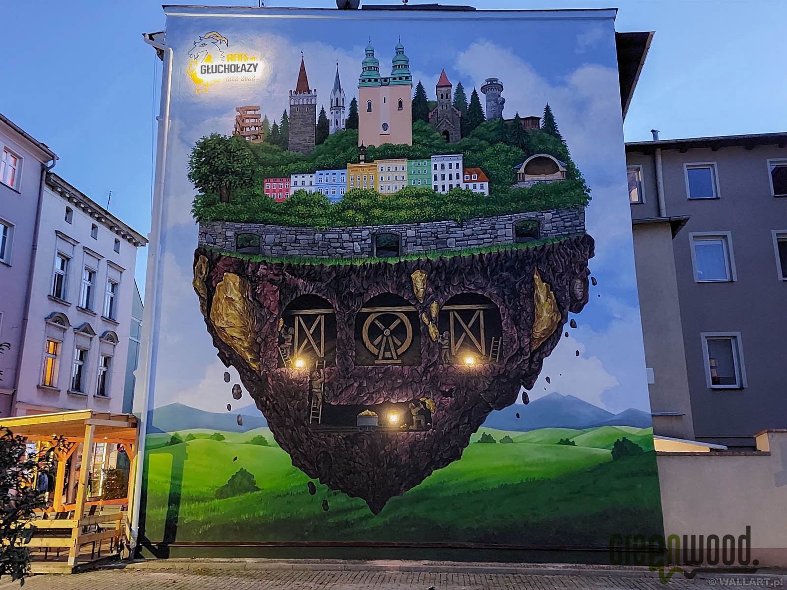 Mural na ścianie budynku przedstawiający fantazyjny krajobraz z lewitującą wyspą, na której znajduje się miasto z wieżami i górniczymi szybami, całość utrzymana w jasnej kolorystyce.