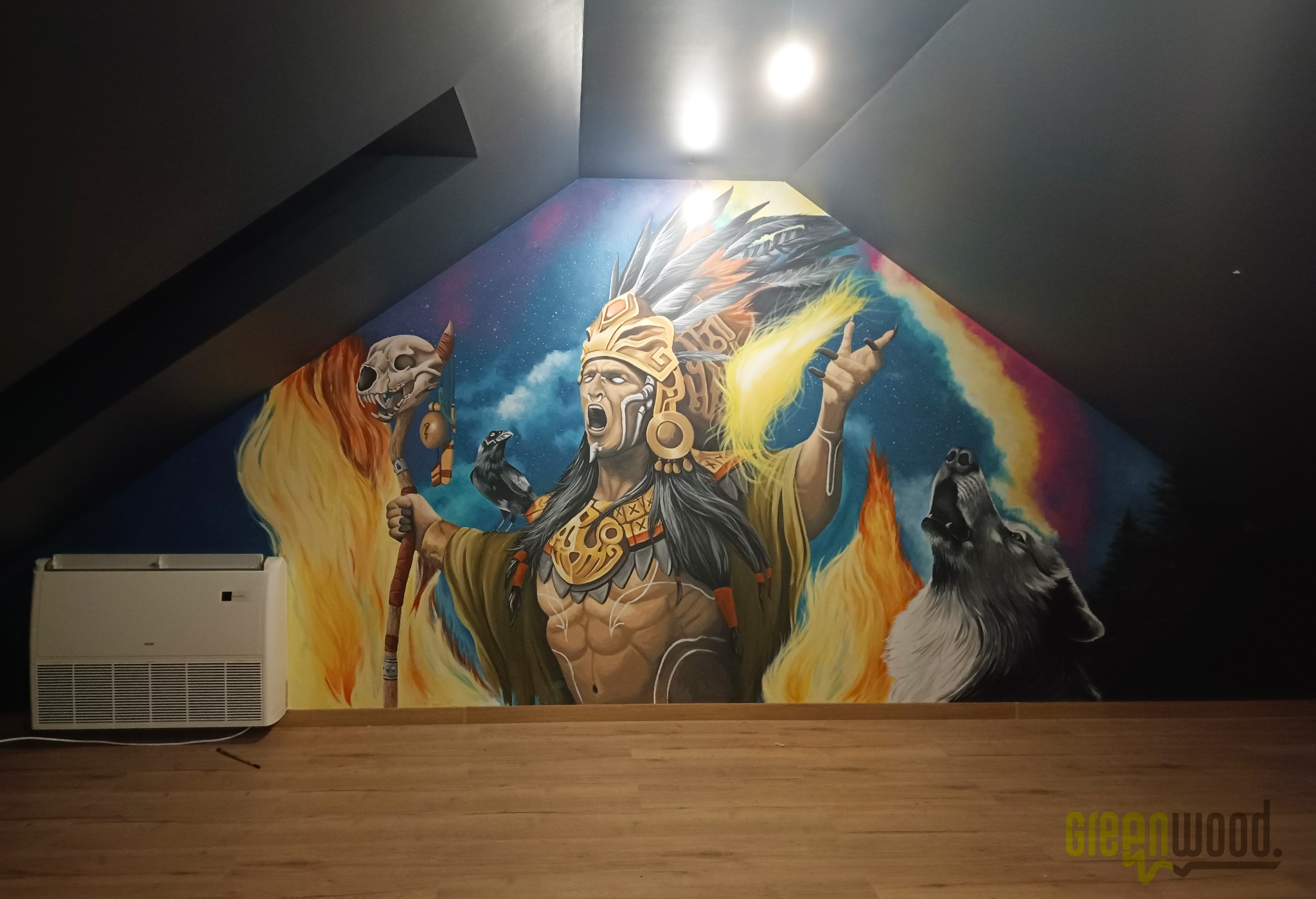 Imponujący mural w pokoju na poddaszu przedstawiający szamana w nakryciu głowy z piór, czaszkę zwierzęcą i wyjącego wilka na tle kosmosu i tęczy, z klimatyzacją i drewnianą podłogą w dolnej części.
