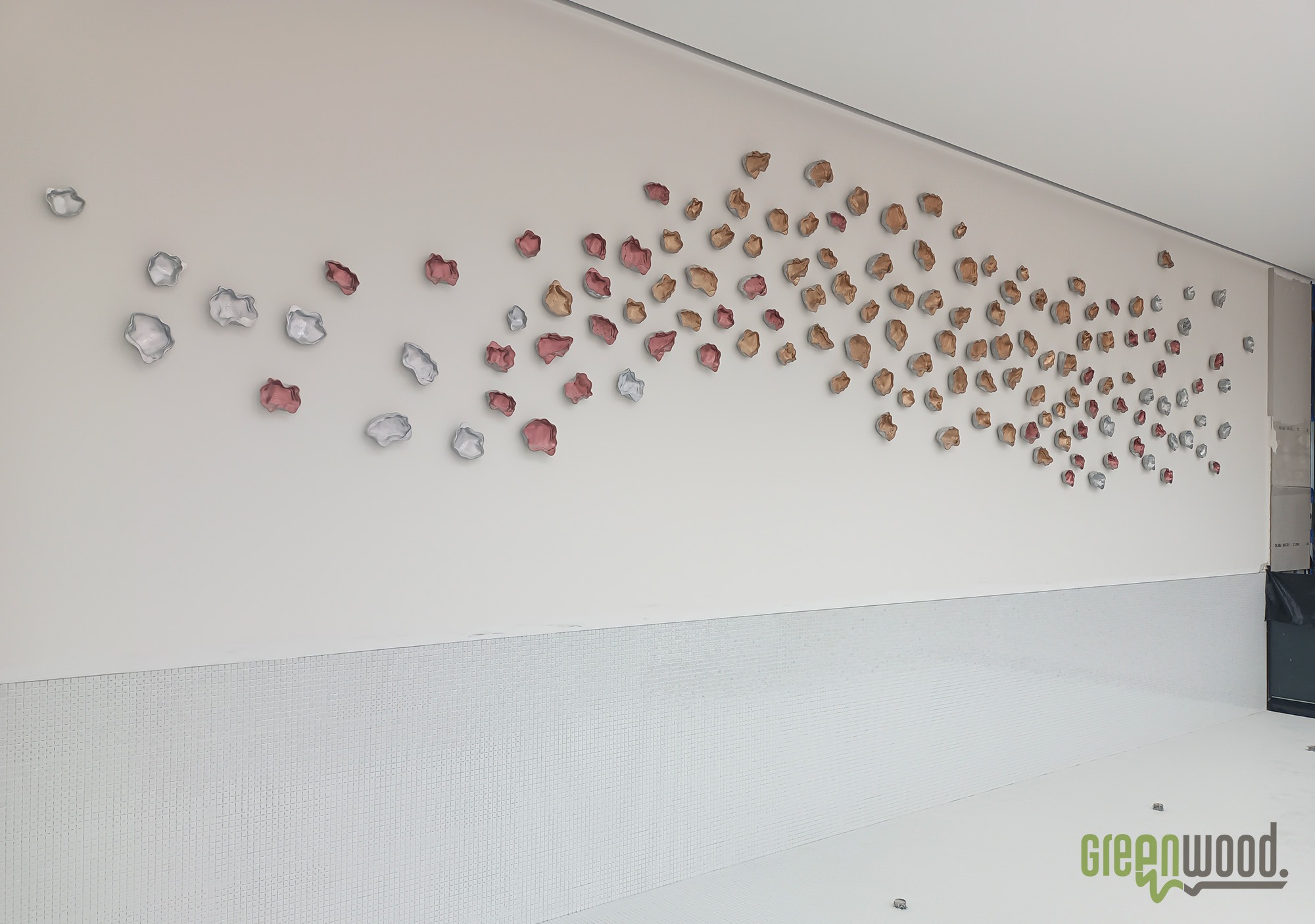 Dekoracyjna instalacja ścienna z trójwymiarowych elementów w kształcie płatków, stopniowo zmieniających kolor od srebrnego i różowego do brązowego, na białej ścianie z dolnym pasem mozaiki.