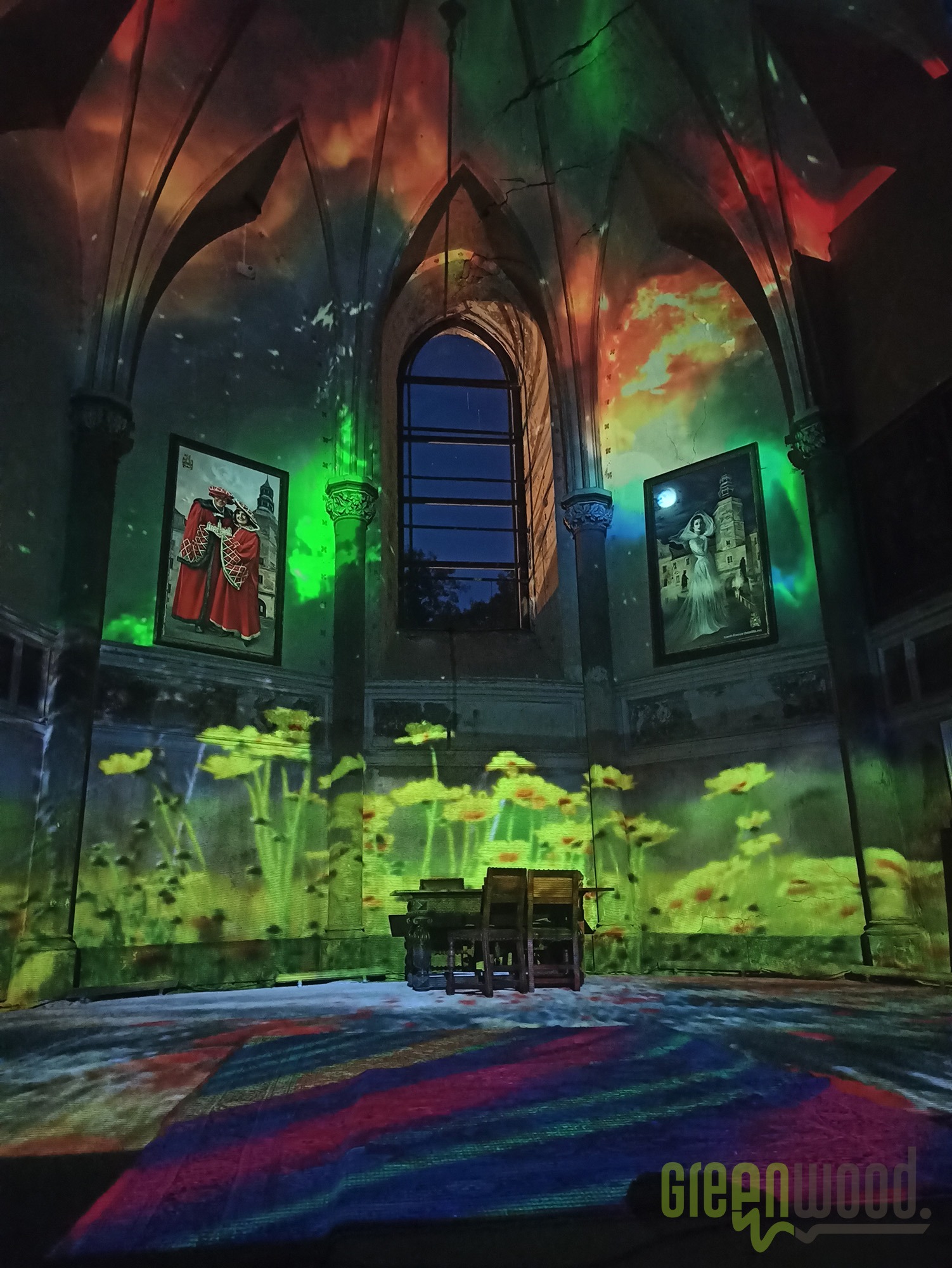 Wnętrze gotyckiej budowli z projekcjami świetlnymi kwiatów i abstrakcyjnych wzorów, obrazy na ścianach podświetlone zielonym światłem, drewniane krzesła w centrum.