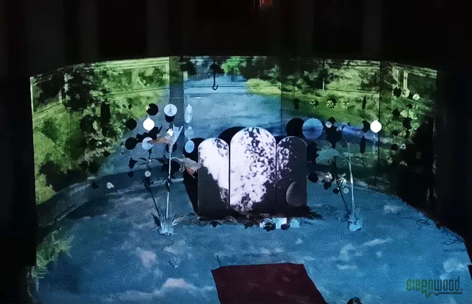 Scenografia teatralna z projekcjami: stylizowany ogród z nagrobkami w tle, oświetlony dekoracyjnymi lampkami i z elementami roślinnymi.