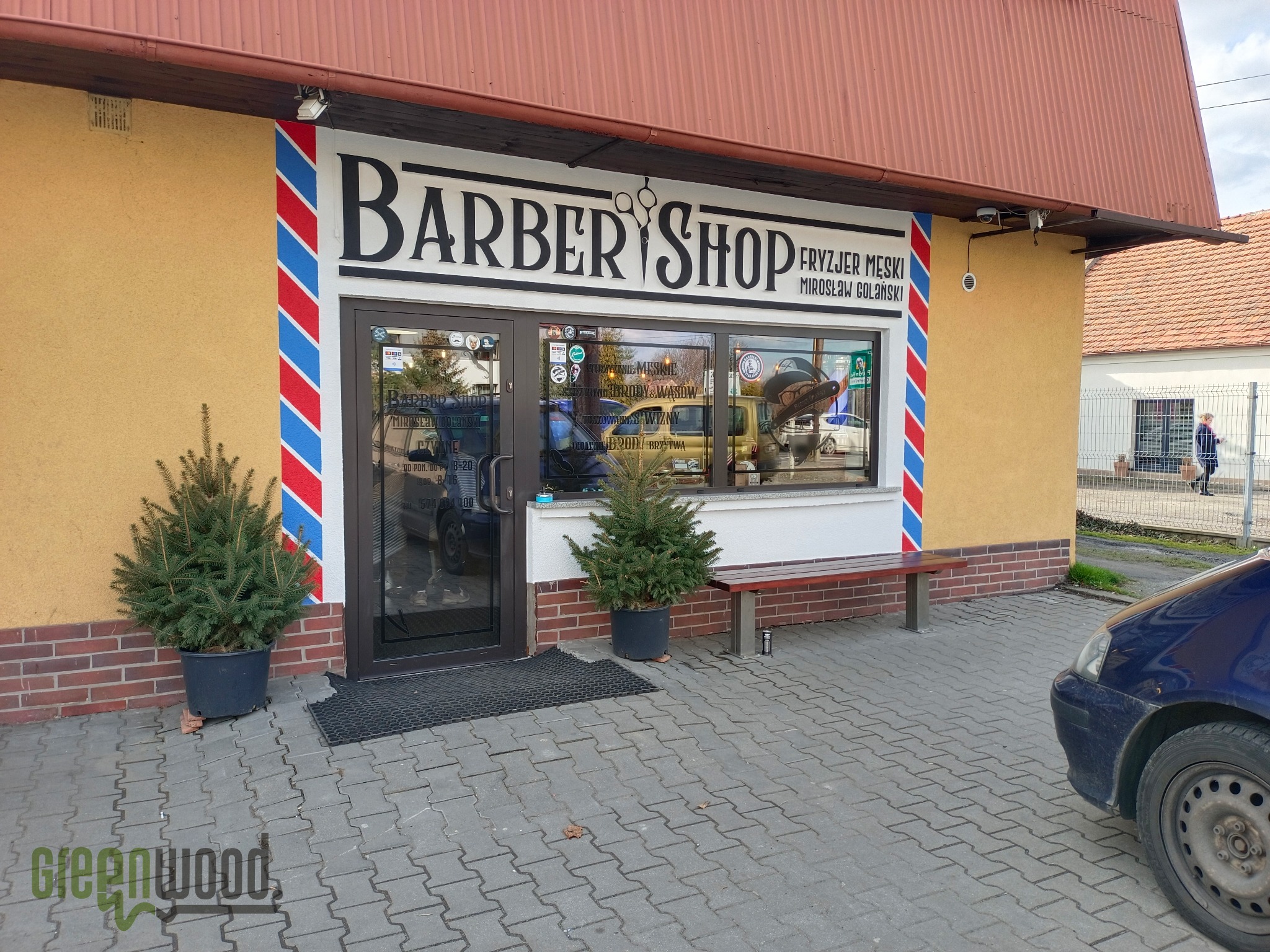 Fasada budynku z szyldem 'Barber Shop Fryzjer Męski Mirosław Golański', ozdobiona czerwono-biało-niebieskimi pasami, z dwoma małymi drzewkami iglastymi w donicach po bokach wejścia i ławką przed...