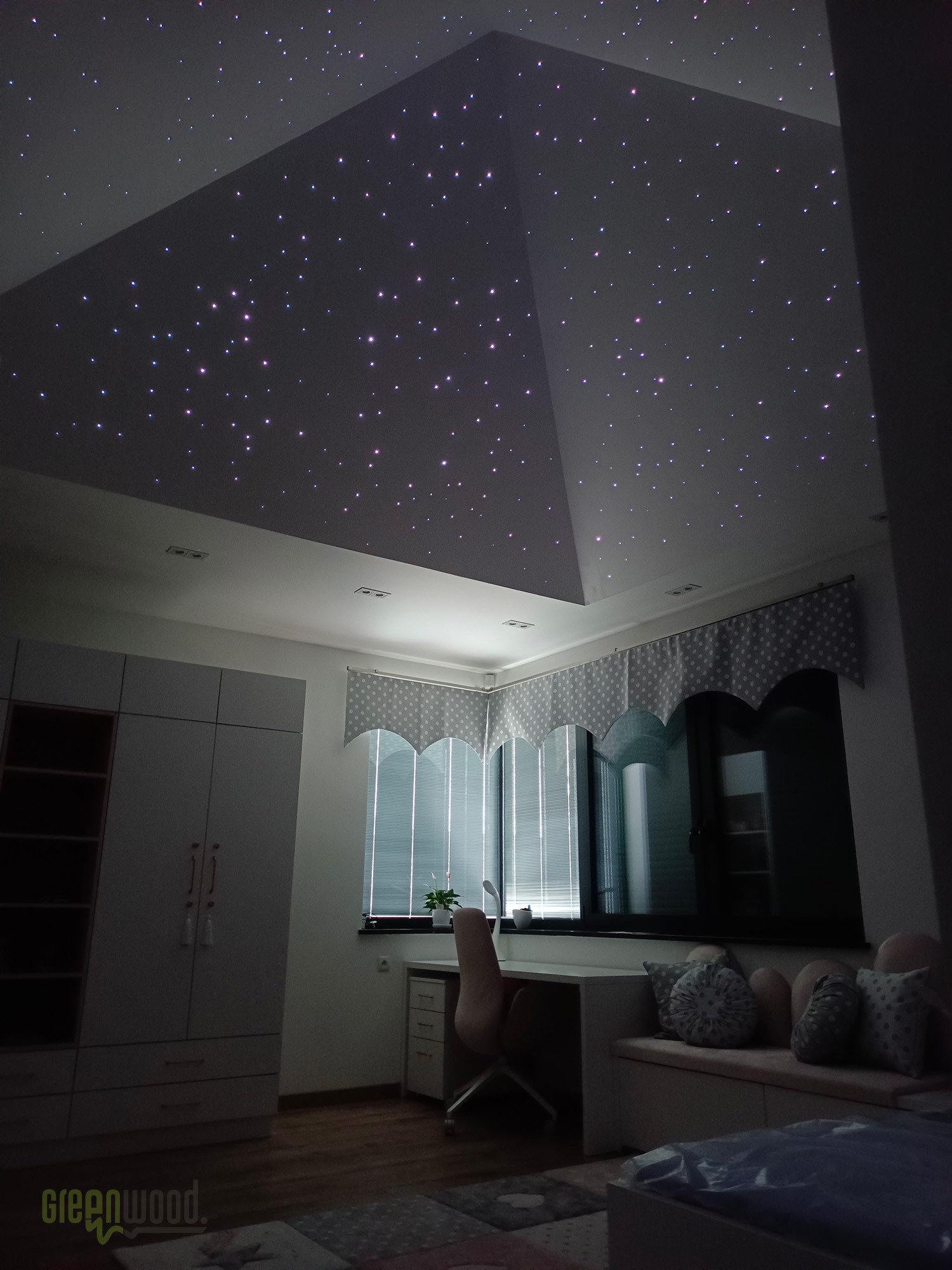 Pokój dziecięcy z oświetleniem sufitowym imitującym rozgwieżdżone niebo, widoczny biały regał, biurko z krzesłem i okno z zasłonami w kropki.