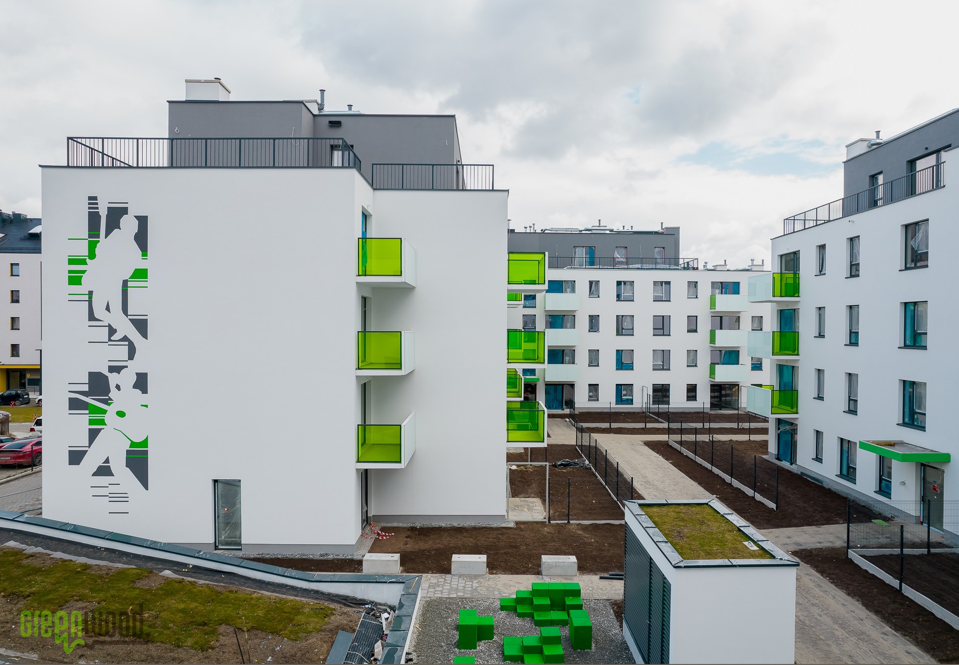 Nowoczesne osiedle z białymi budynkami, zielonymi balkonami i abstrakcyjną grafiką na ścianie, widok z góry na dziedziniec z elementami małej architektury.