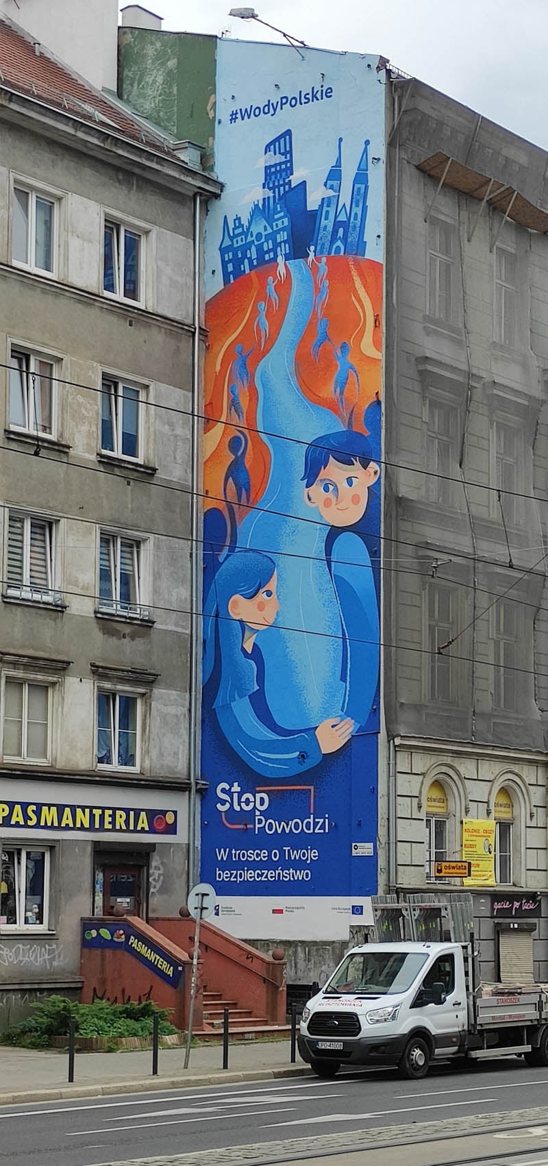 Mural na ścianie budynku przedstawiający stylizowaną parę w niebieskich odcieniach, otoczoną motywem rzeki i sylwetkami ludzi, z hasłem 'Stop Powodzi', widoczny fragment ulicy i zaparkowany biały...