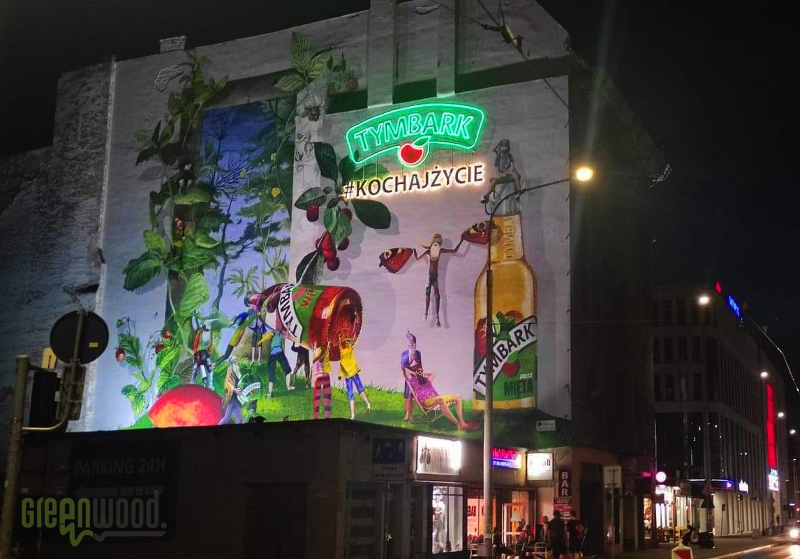 Iluminowany mural na ścianie budynku przedstawiający motywy roślinne, postacie i logo marki napoju, wykonany w żywych kolorach i widoczny nocą.