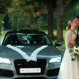 Pocałunek pary młodej na tle udekorowanego samochodu Audi RS7 z białą wstążką i kwiatami, droga obsadzona drzewami w tle.