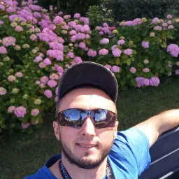 Mężczyzna w czapce i okularach przeciwsłonecznych siedzi na ławce na tle krzewów kwitnących na różowo w parku, robiąc sobie selfie.