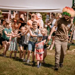 Animator w kolorowej peruce prowadzi korowód dzieci po trawie podczas imprezy plenerowej.
