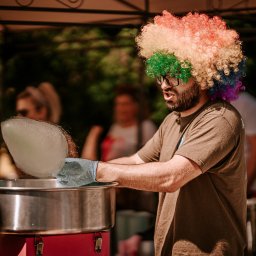 Amigol Krzysztof Jurgielewicz - Mężczyzna w okularach i kolorowej peruce robi watę cukrową na metalowej maszynie, w tle widoczne osoby.
