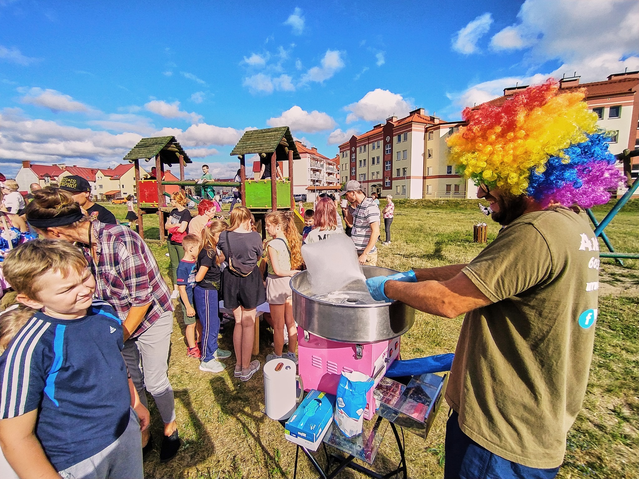 Mężczyzna w kolorowej peruce przygotowuje watę cukrową na różowym urządzeniu podczas imprezy plenerowej dla dzieci, widoczny plac zabaw i budynki w tle.