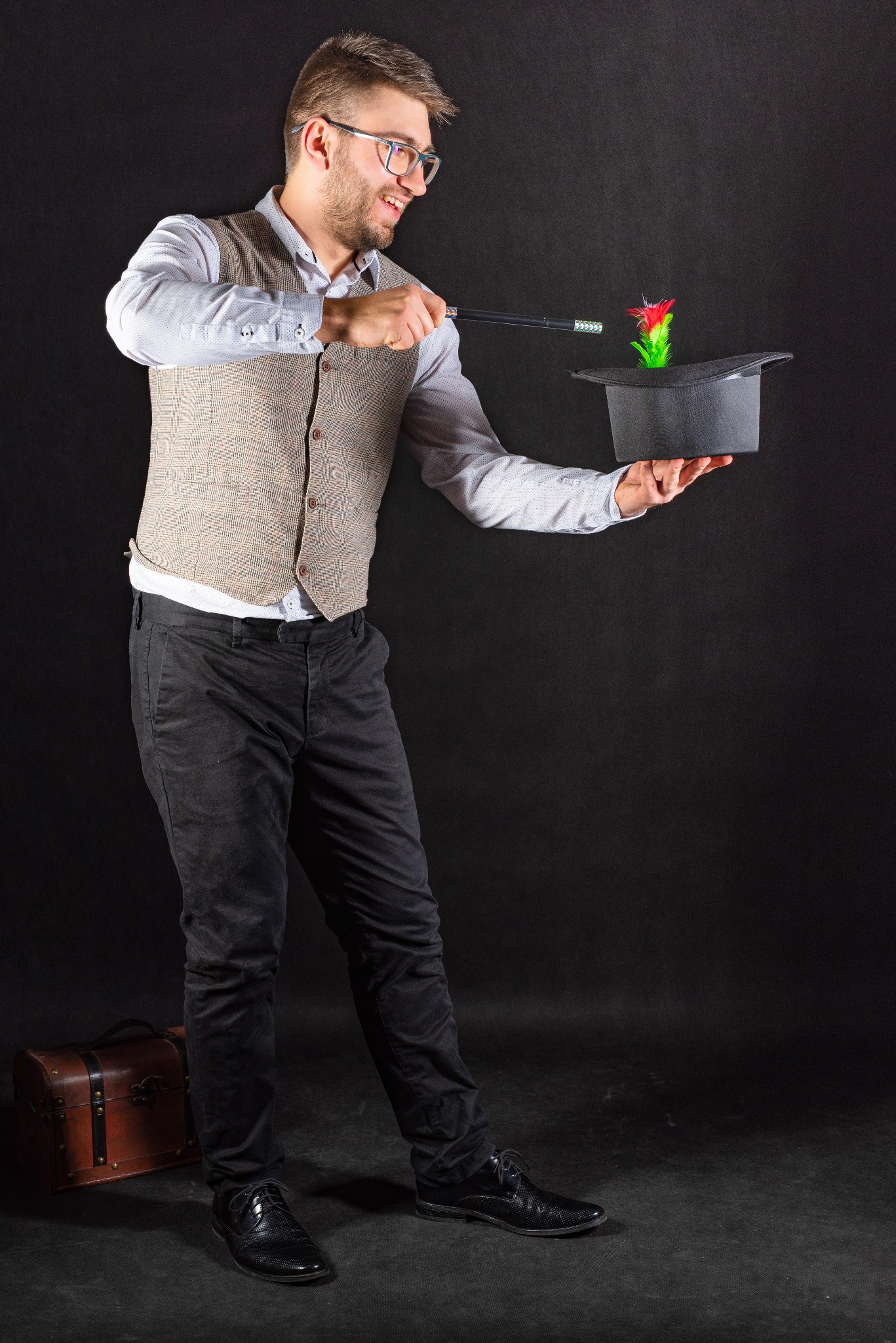 Młody mężczyzna w okularach, kamizelce i koszuli, uśmiechnięty, trzyma czarny cylinder, z którego wystaje czerwono-zielony rekwizyt przypominający kwiat, machając nad nim czarną różdżką; w tle...