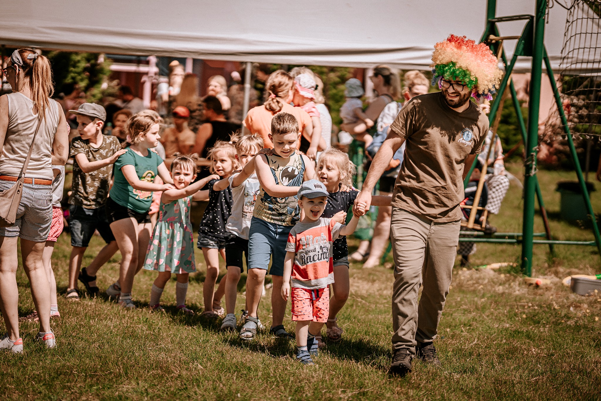 Animator w kolorowej peruce prowadzi korowód dzieci po trawie podczas imprezy plenerowej.