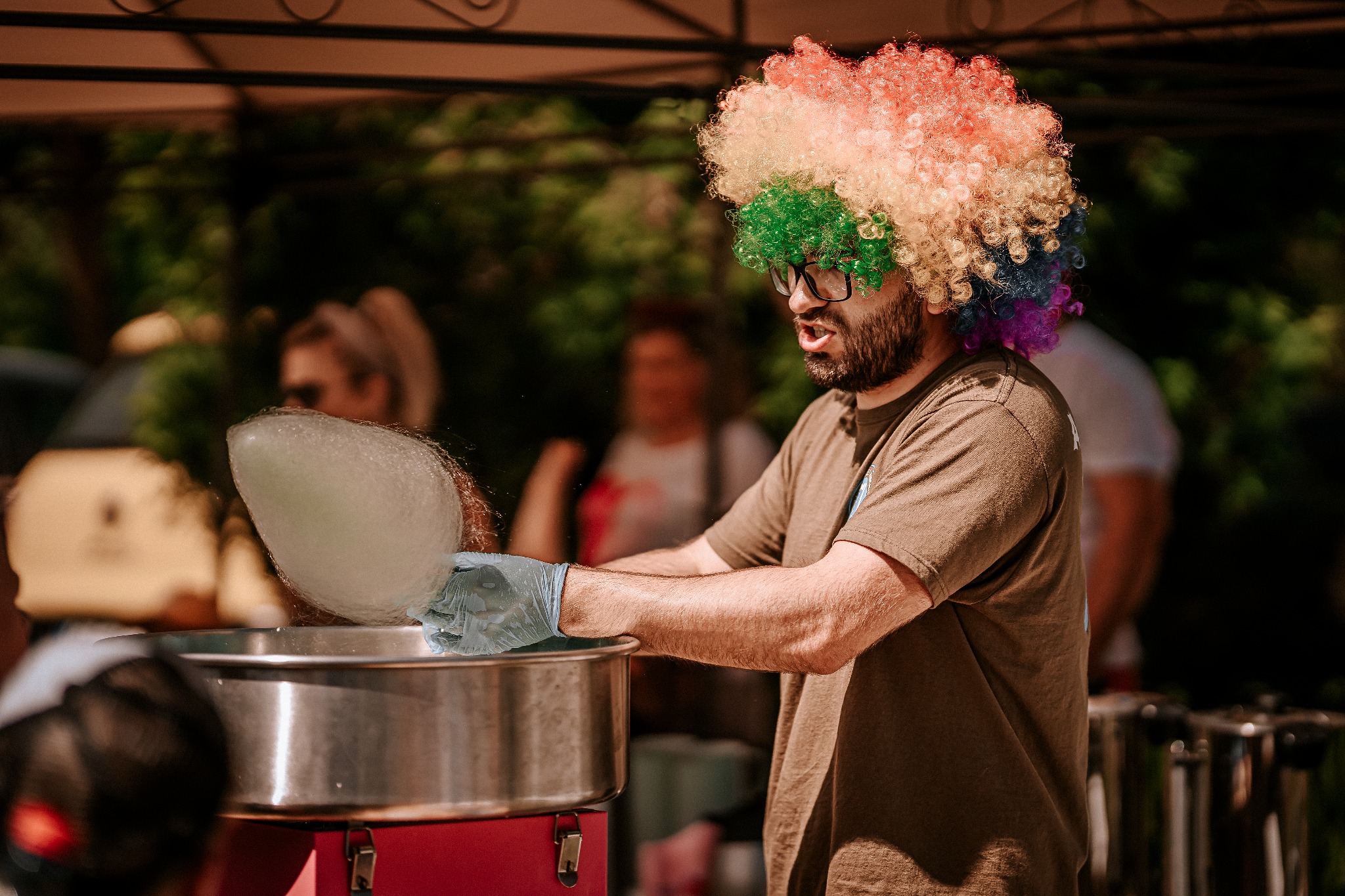Mężczyzna w okularach i kolorowej peruce robi watę cukrową na metalowej maszynie, w tle widoczne osoby.